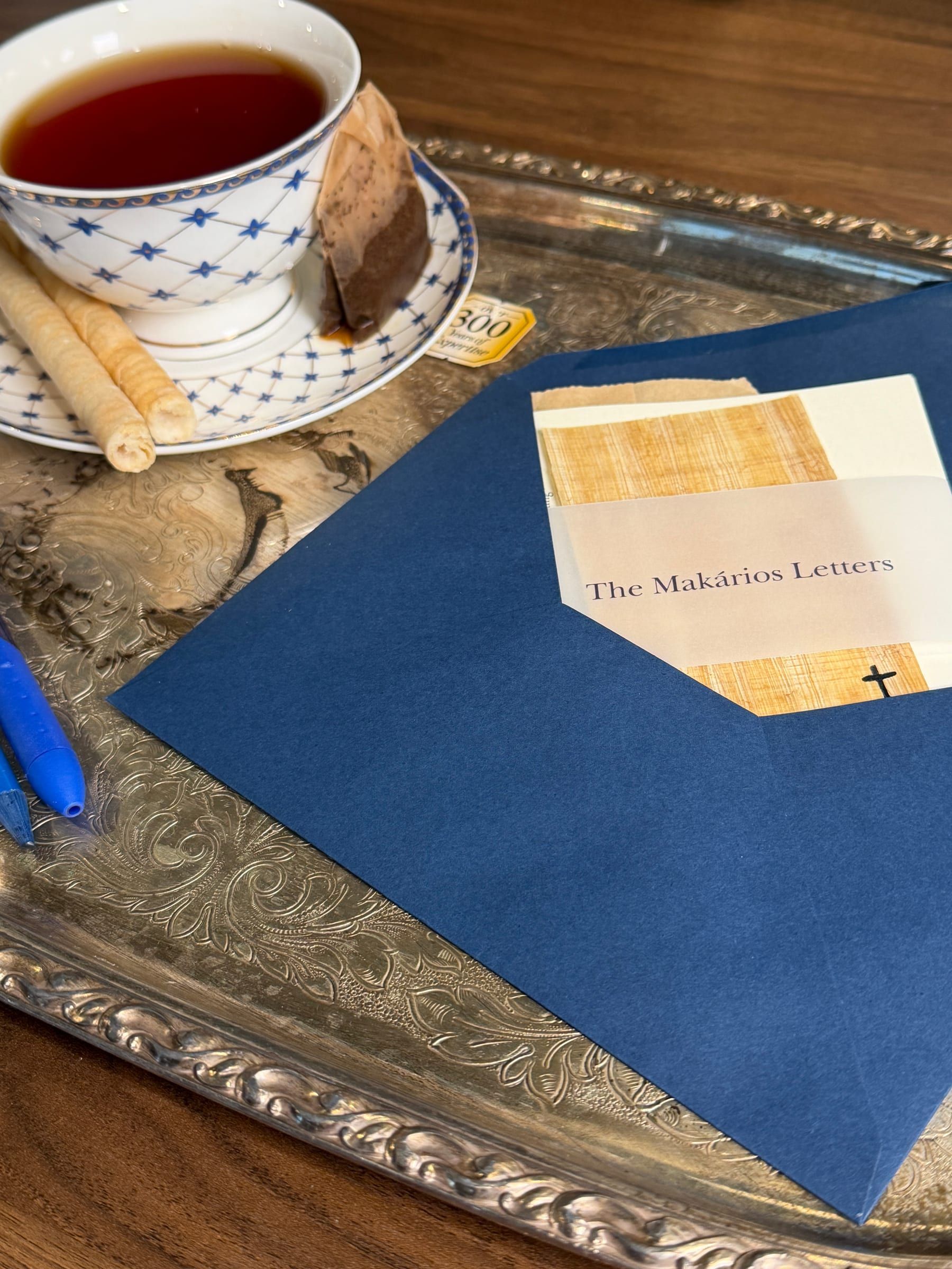 The Makários Letters in a vellum band over papyrus, tucked in a navy envelope beside a cup of tea on a silver tray.