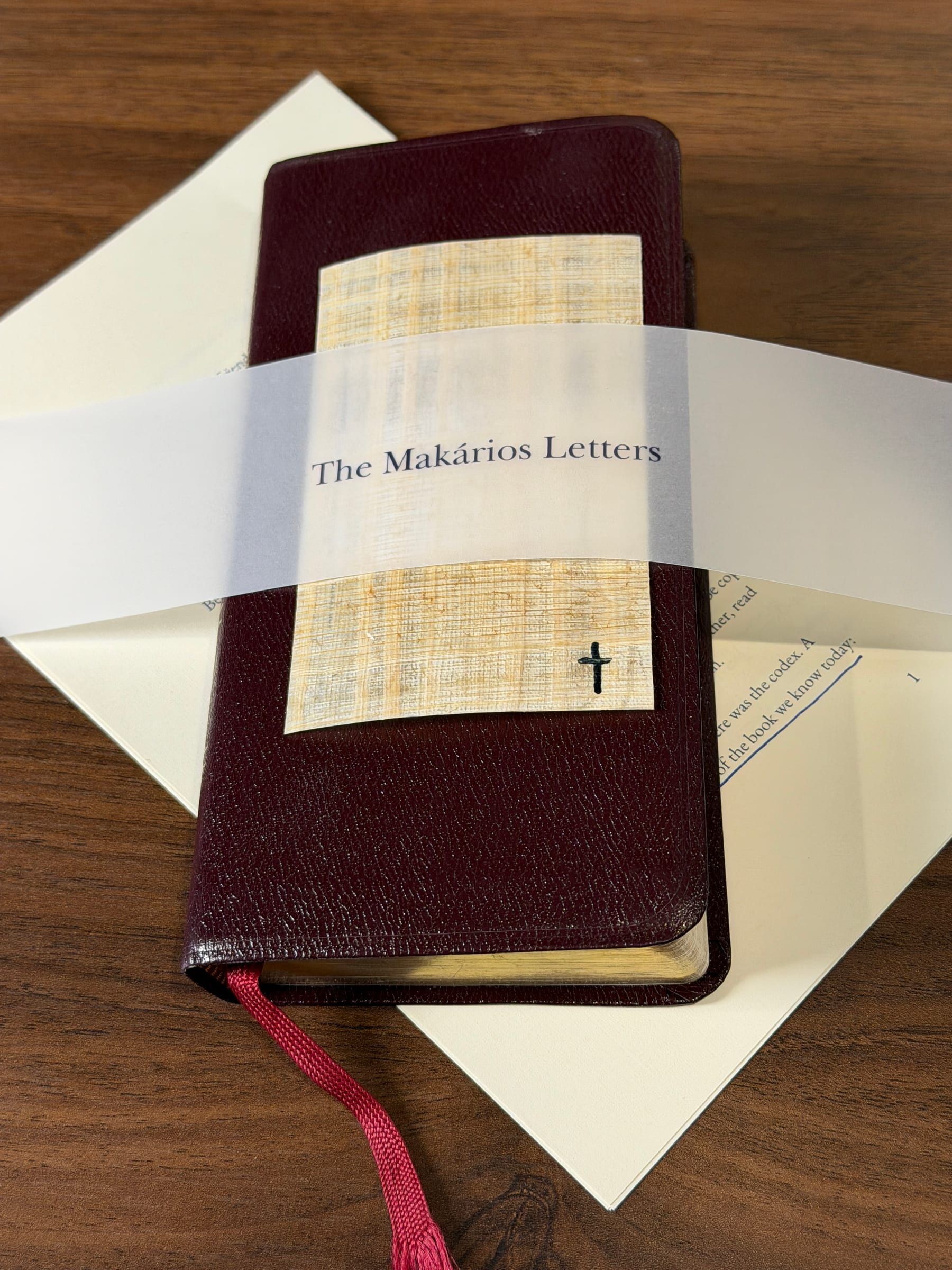 A vellum band printed “The Makários Letters” wraps a papyrus card with a small cross, on a burgundy leather Bible.