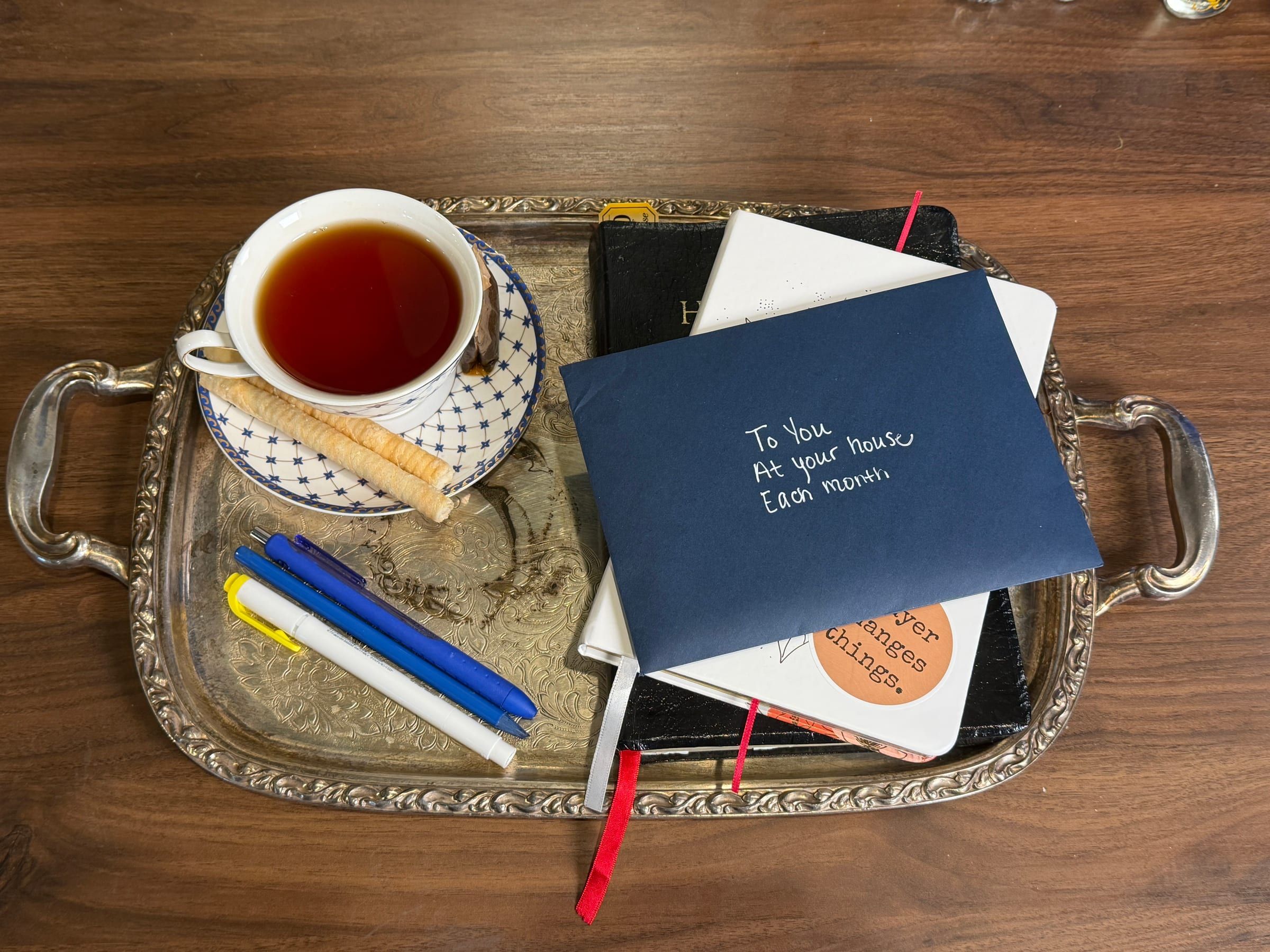 From above: a navy envelope addressed “To You, At your house, Each month” on a Bible and a journal, with tea and pens on a silver tray.