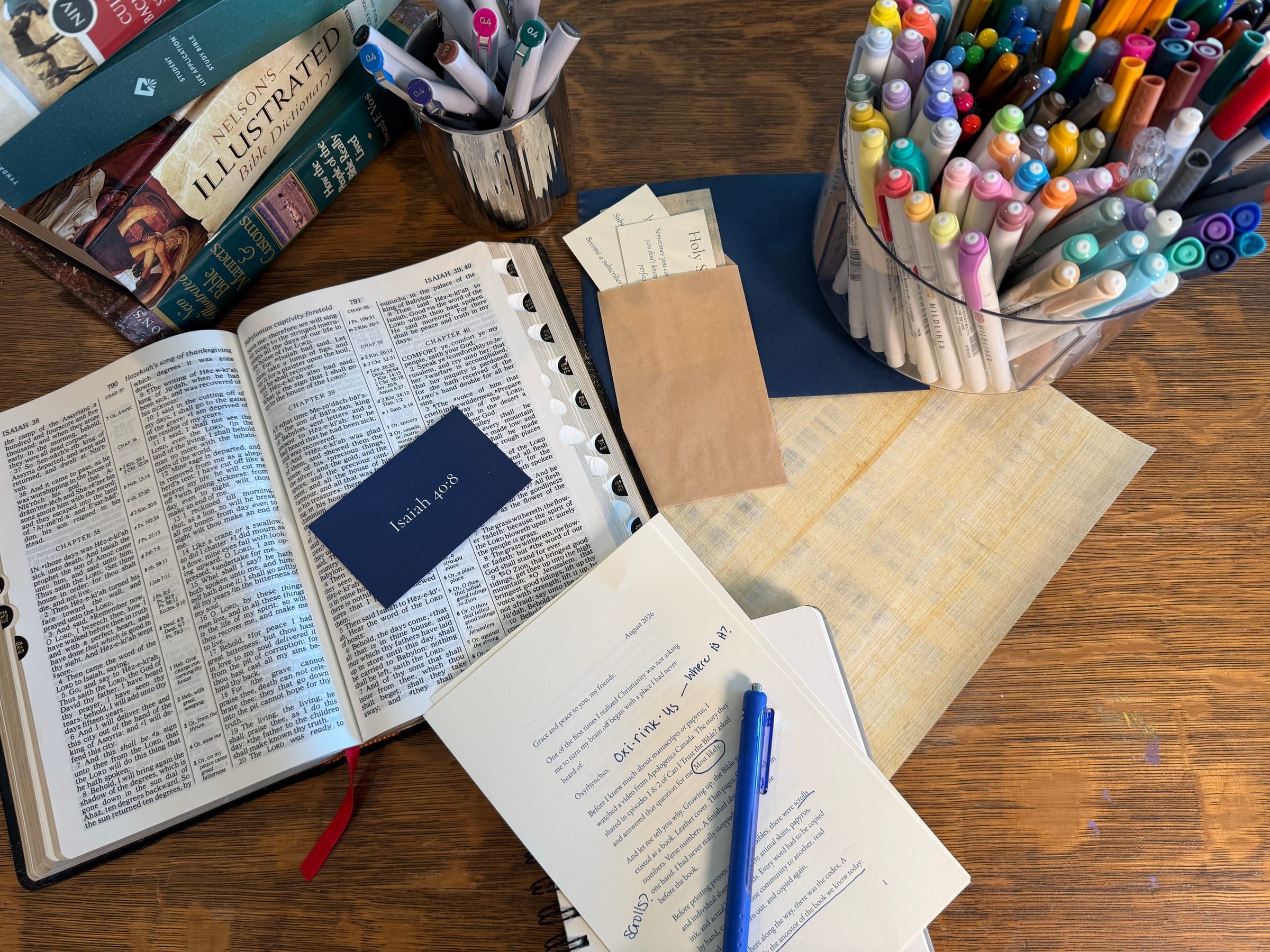 From above: an open Bible with an Isaiah 40:8 card, the marked-up August letter, a kraft envelope of cards, papyrus, and jars of colored pens.
